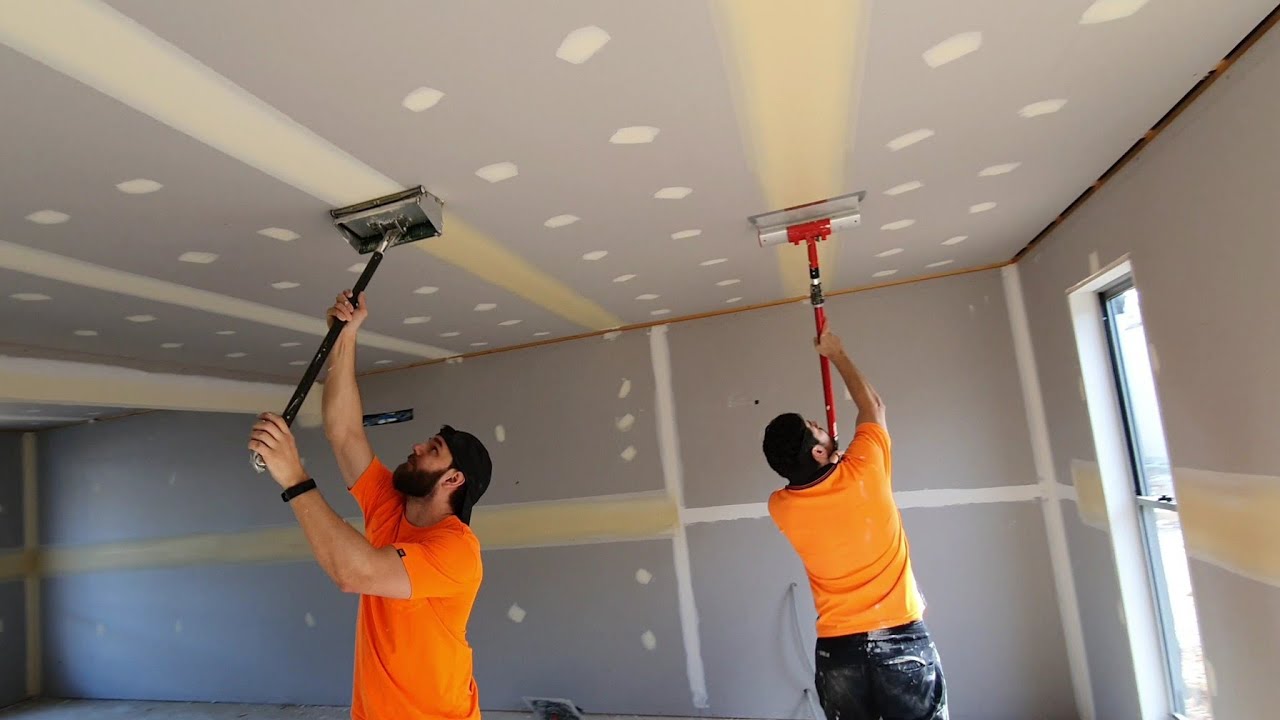 Two men in orange shirts sanding drywall on a ceiling of an unfinished room.