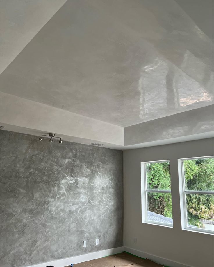 Empty room with polished concrete accent wall, glossy ceiling, two windows showing green trees outside.