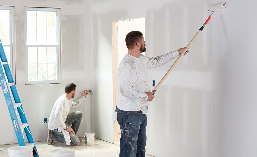Two men in white shirts painting drywall inside a room under renovation.