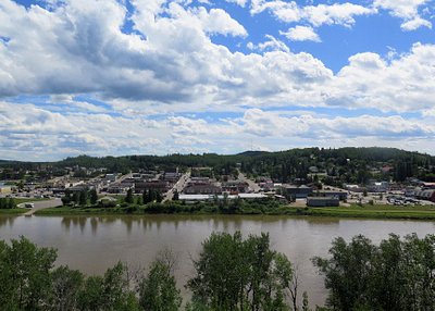 View of a town beside a river with trees in foreground and hills under a blue sky with clouds.