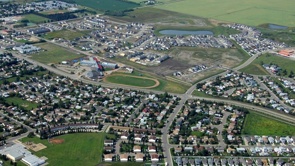 Aerial view of suburban neighborhood with houses, roads, a school, sports fields, and a small pond.