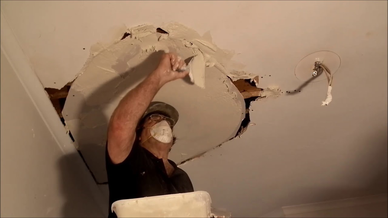 Man wearing a mask and cap repairs a ceiling hole by applying plaster with a trowel.