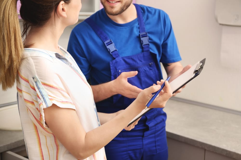 Woman and technician in blue overalls discussing while holding clipboard and pen in kitchen.