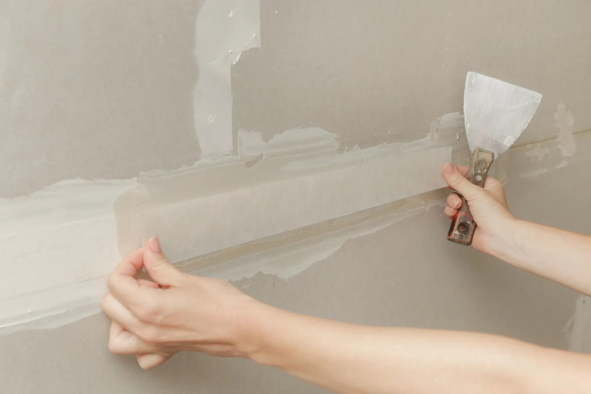 Person applying drywall tape to a joint on a wall using a putty knife.