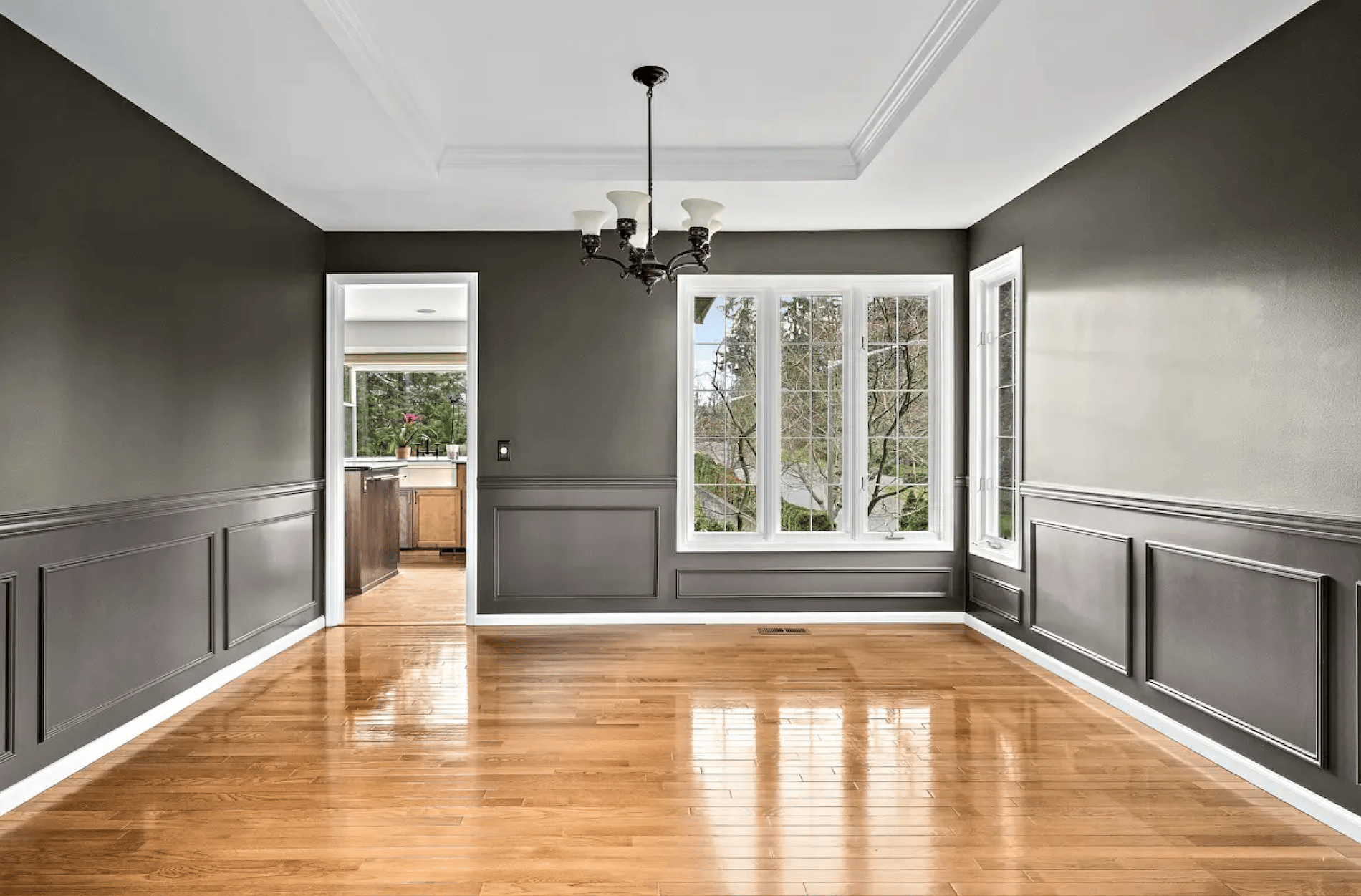 Empty dining room with dark gray walls, hardwood floor, chandelier, and view into kitchen through doorway.