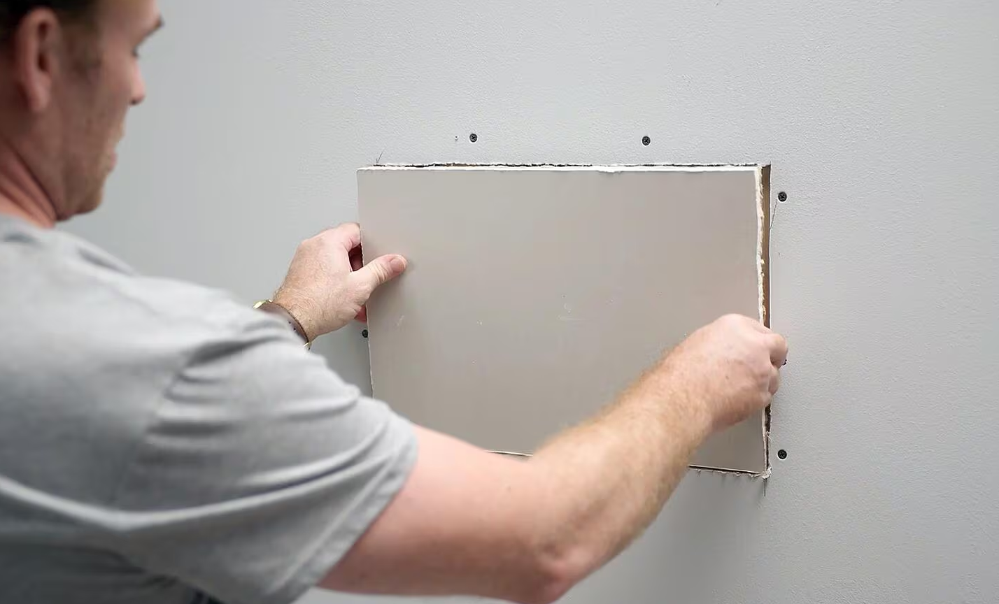 Man placing a drywall patch into a rectangular cutout in a wall.