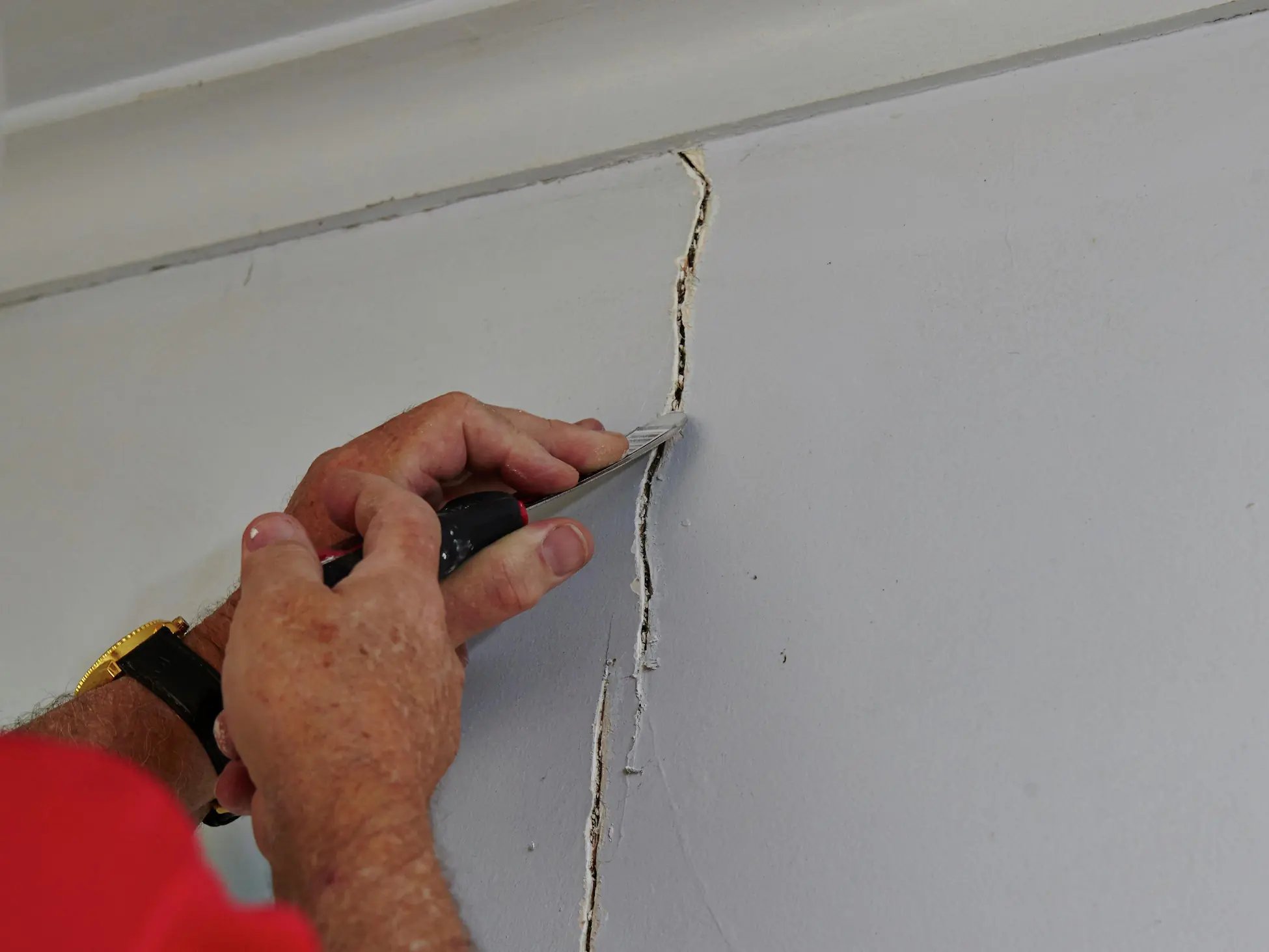 Person using a utility knife to widen a crack on a white wall for repair.