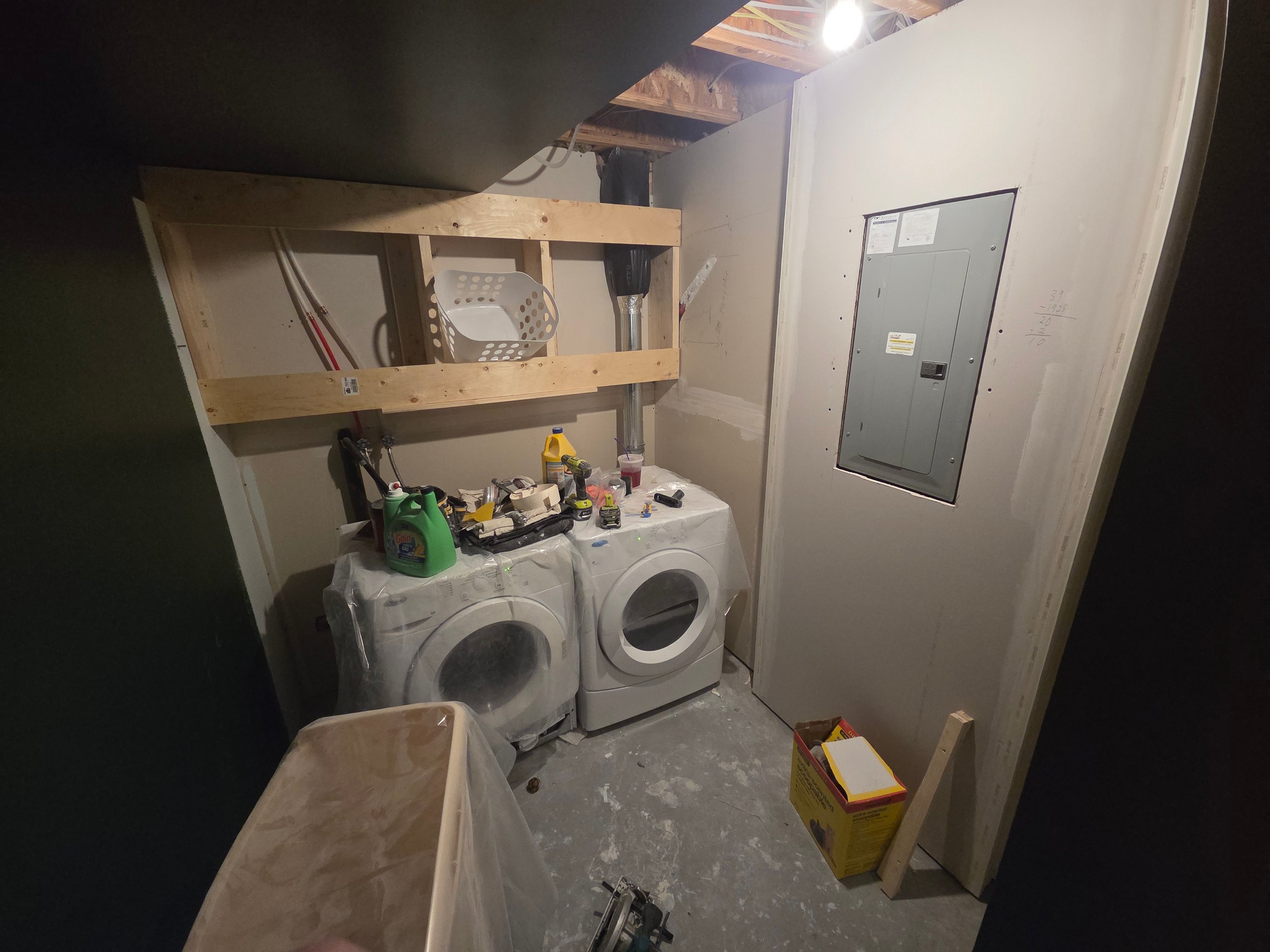 Laundry area under construction with washer, dryer, wooden shelf frame, and electrical panel on wall.