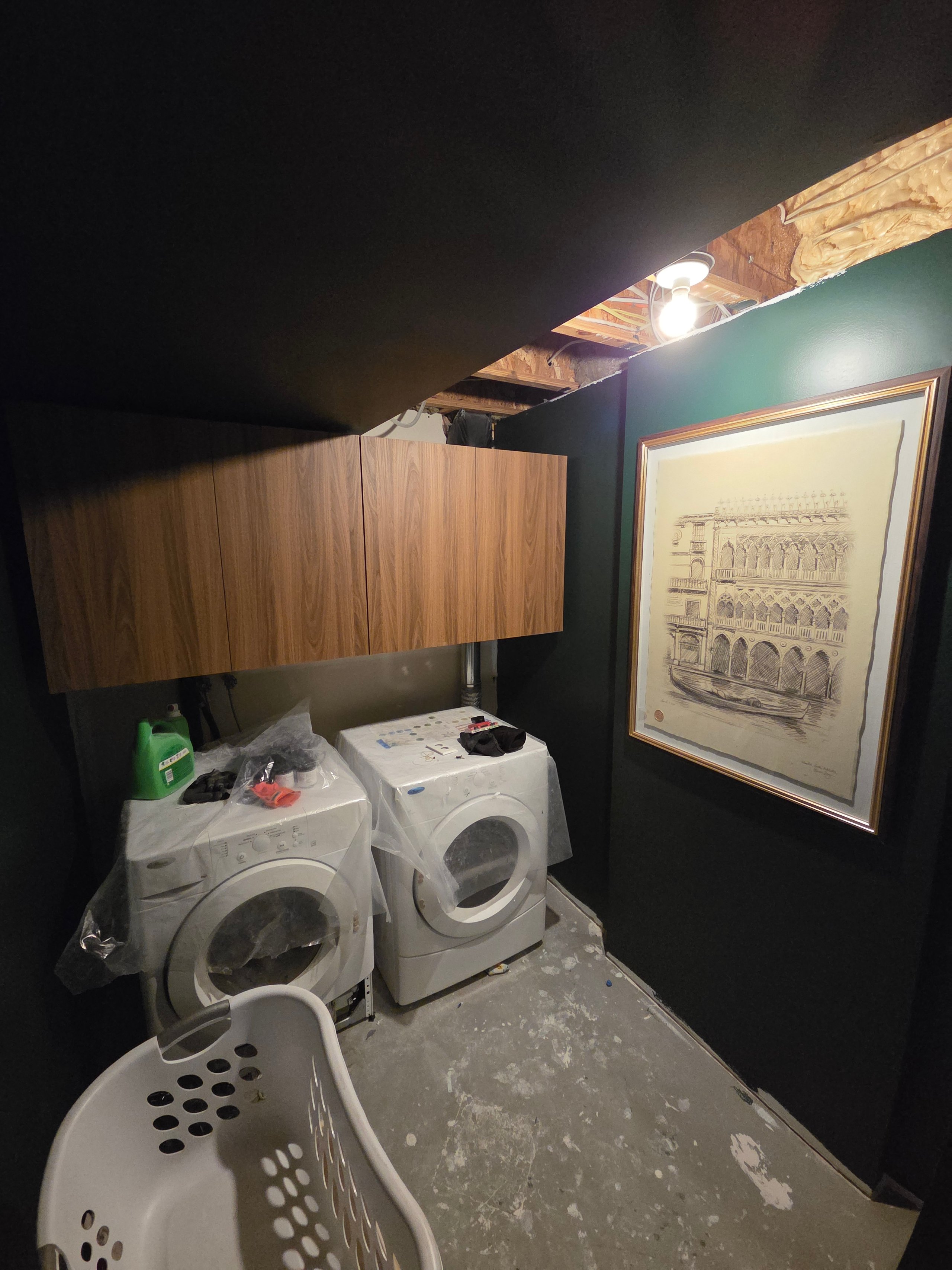 Basement laundry room with washer, dryer, laundry basket, wood cabinets, and framed wall art.