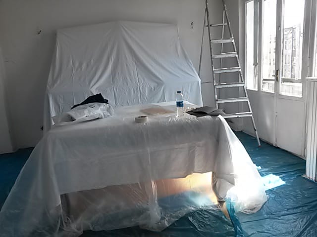 Room under renovation with ladders, furniture and floor covered in protective plastic and blue sheets.