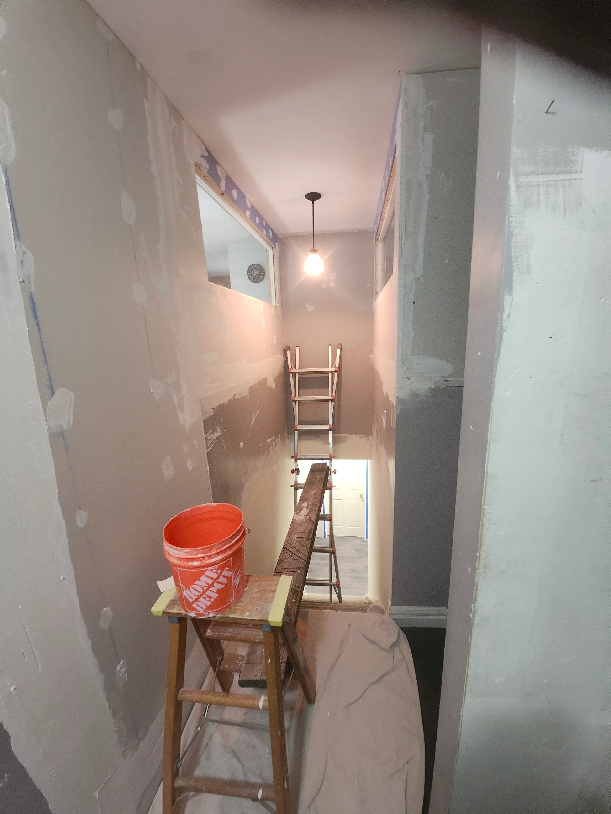 Narrow stairwell under renovation with a ladder, plank, paint bucket, and a hanging light bulb.