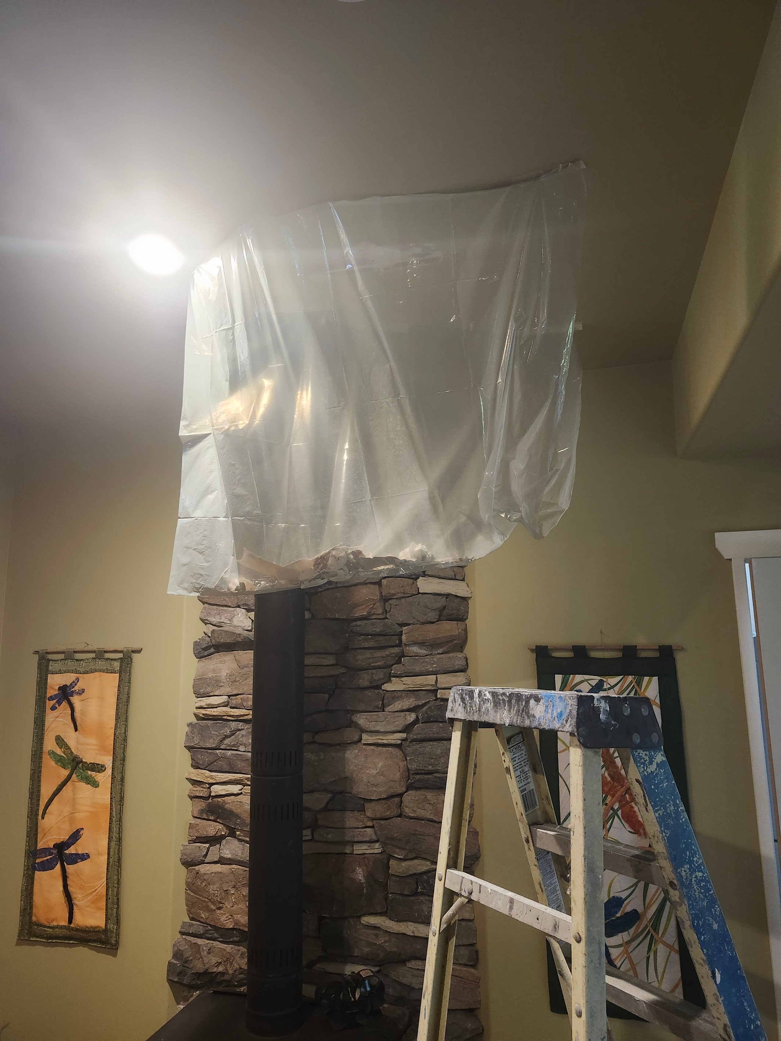 Plastic sheet hanging from ceiling above stone fireplace vent with a paint-splattered ladder in front.