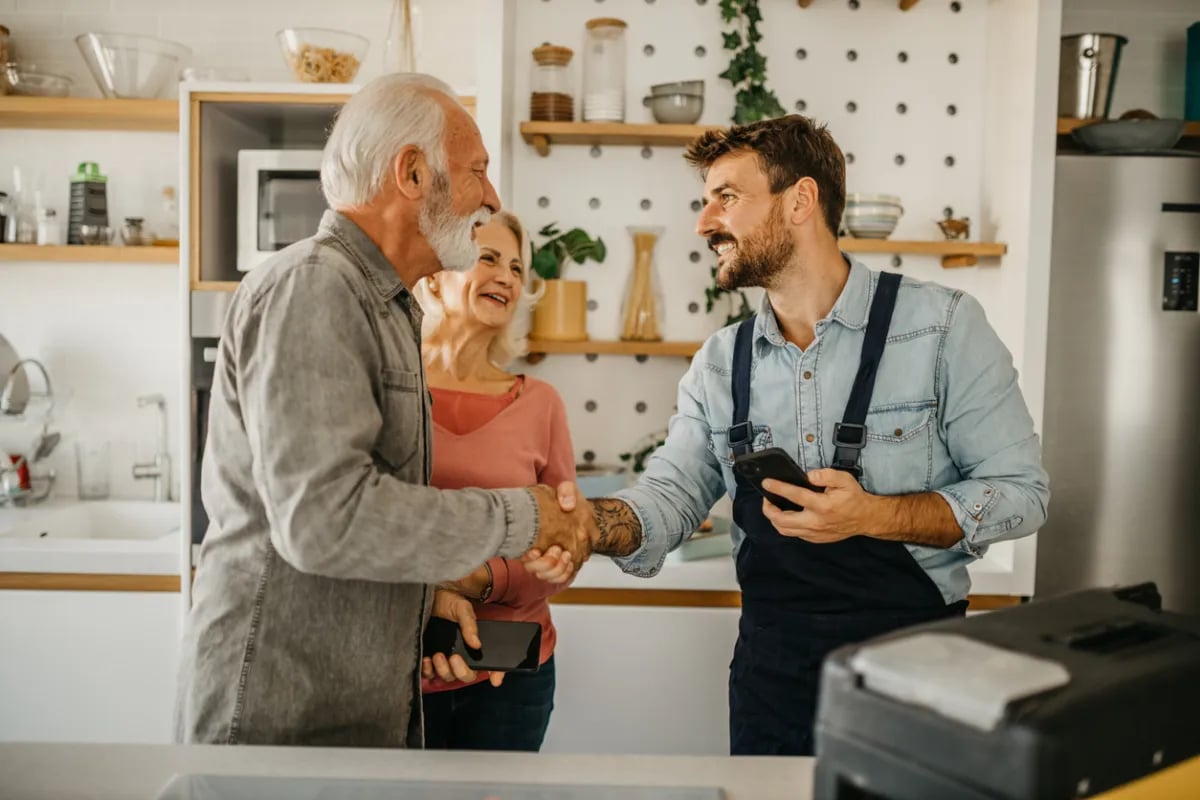 Older couple shaking hands with a younger man in an apron inside a bright kitchen.