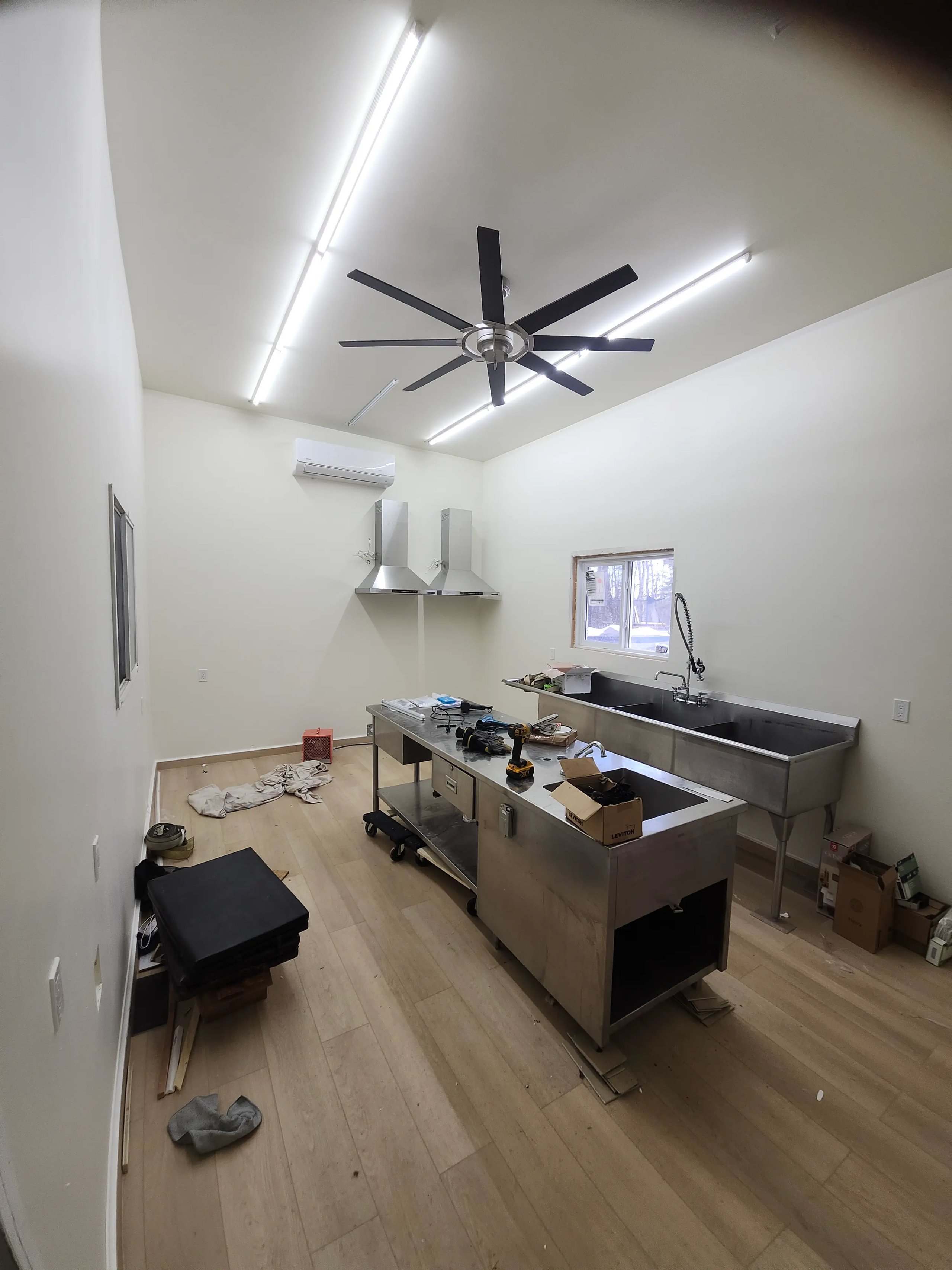 Minimalist kitchen with stainless steel island, dual sinks, large ceiling fan, and scattered tools and boxes.