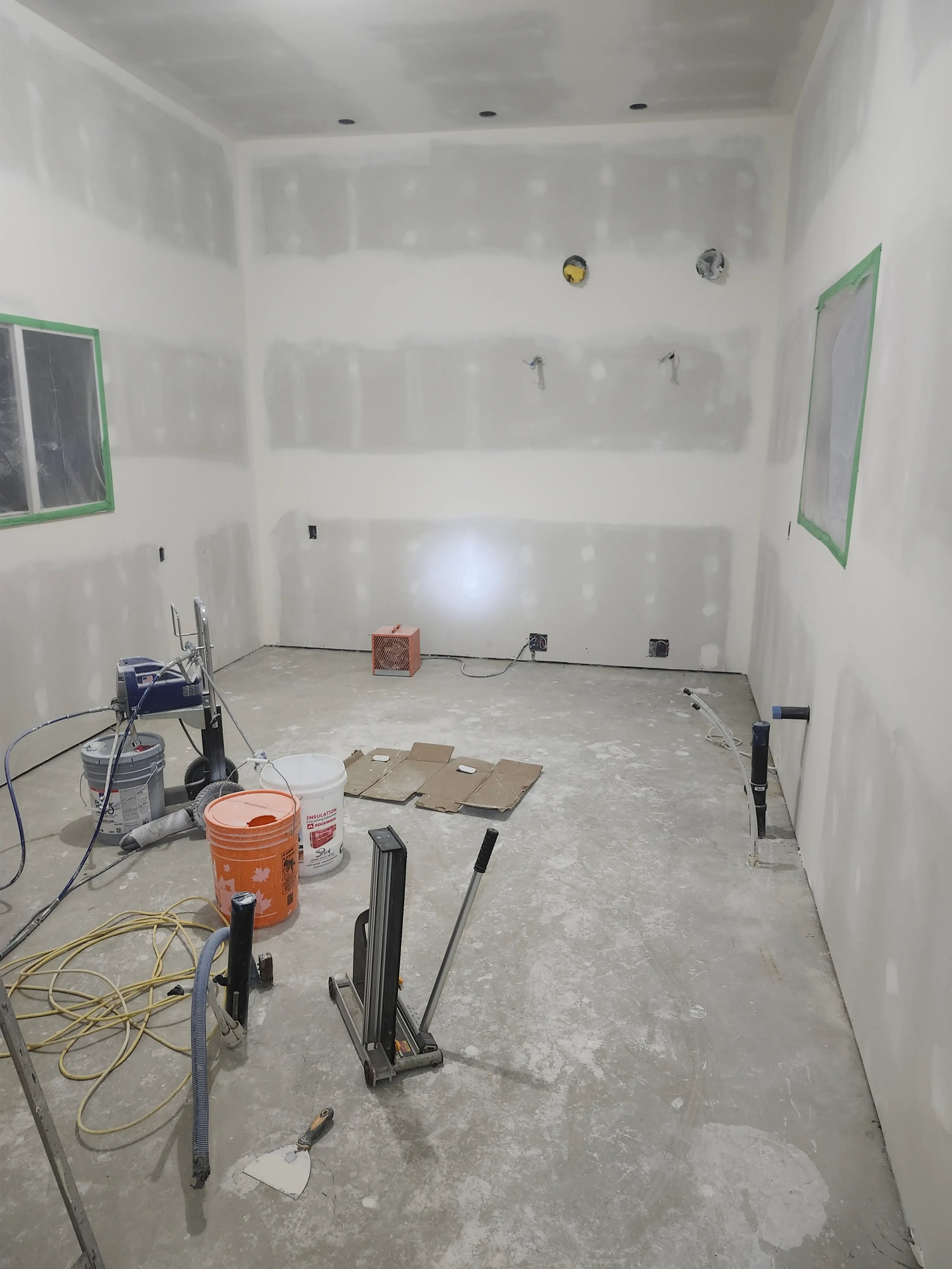 Room under construction with drywall, tools, buckets, wires, and taped windows ready for finishing.