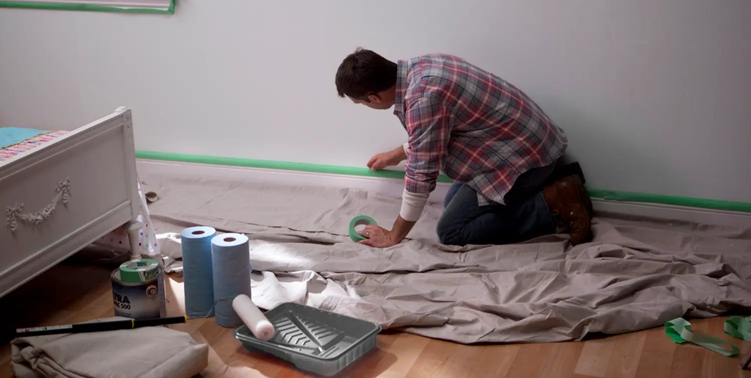 Man kneeling on drop cloth applying green painter's tape to a white wall baseboard.